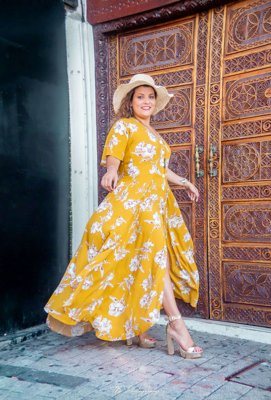 Femme souriante en robe longue jaune fleuri, posant devant une porte en bois ornée.