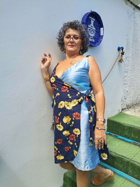 Femme en robe portefeuille florale bleue et noire, posant devant un mur blanc, style décontracté et élégant.