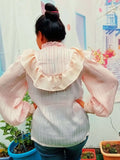 Femme portant une blouse Fulla avec col montant froncé et manches Bishop, vue de dos, jardin en arrière-plan.