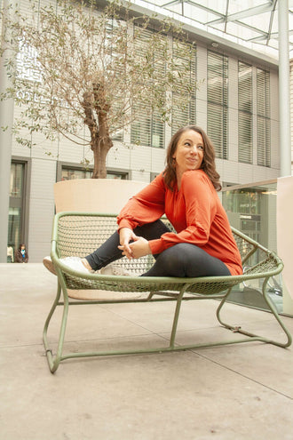 Femme assise sur un banc en métal dans un espace urbain moderne, portant un pull orange vif.