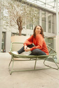 Femme assise sur un banc en métal dans un espace urbain moderne, portant un pull orange vif.