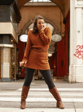 Femme portant un pull long marron avec ceinture, debout dans une rue couverte.
