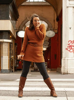 Femme portant un pull long marron avec ceinture, debout dans une rue couverte.