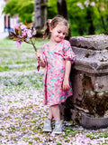 Petite fille souriante en robe fleurie tenant des fleurs rose, posant à l'extérieur sur un chemin jonché de pétales.