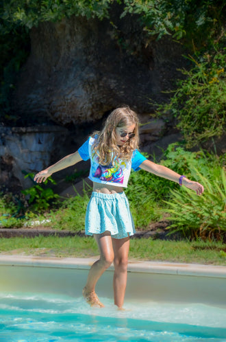 Young girl in a colorful outfit, playfully walking in shallow water at a sunny outdoor setting.