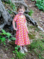 Enfant portant une robe colorée en promenade dans la forêt, souriante près des racines d'un arbre.