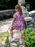 Enfant portant le Patron PDF AMARENA, robe courte fleurie en coton rose et violet, vue de dos dans un jardin