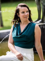 Femme portant la Blouse Bénitier PINA COLADA en satin bleu canard, col bénitier, au bord de l'eau en extérieur
