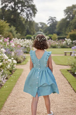 Fille portant le Patron PDF Robe Angélico du 2 au 14 ans, vue de dos en coton bleu ciel, allée de jardin fleuri