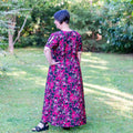 Femme portant le Patron PDF Robe longue Pomme Gala du 34 au 56, manches courtes, fond fleuri, debout dans un jardin verdoyant.