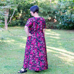 Femme portant le Patron PDF Robe longue Pomme Gala du 34 au 56, manches courtes, fond fleuri, debout dans un jardin verdoyant.