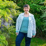 Femme souriante portant une chemise fleurie et un haut turquoise, posant dans un jardin verdoyant.