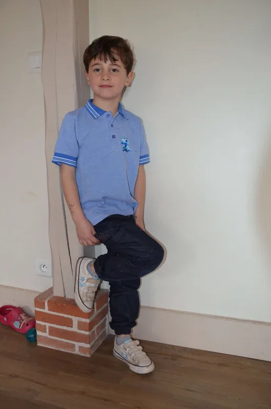 Enfant portant un Polo MARCOLOPO bleu, manches courtes, devant un mur. Patron de couture PDF à télécharger disponible.