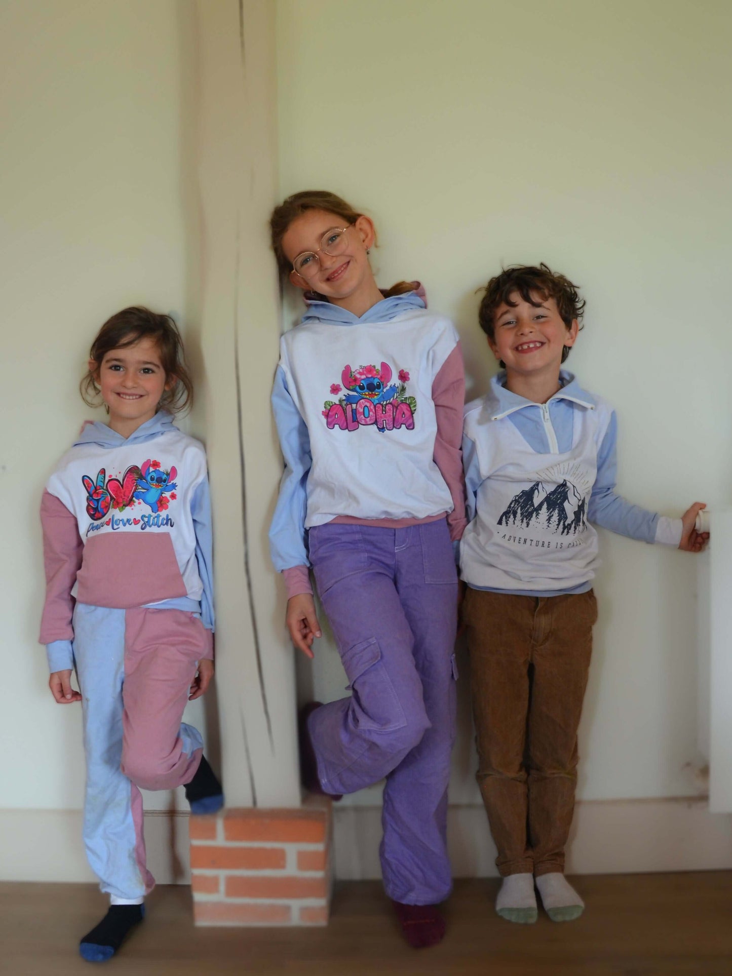 Trois enfants souriants portant des pulls colorés dans une maison.