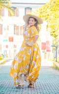 Femme souriante portant une robe longue jaune floral avec chapeau, posant en plein air, ambiance estivale.
