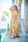 Femme portant une robe longue jaune fleuri avec des manches papillon, posant sur une rue ensoleillée.