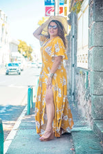 Femme portant une robe longue jaune fleuri avec des manches papillon, posant sur une rue ensoleillée.