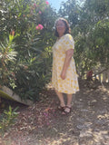 Femme portant une robe jaune à imprimés floraux, se tenant sous un arbre ensoleillé.