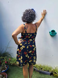Femme portant la robe Ostara à bretelles, montrant un ourlet arrondi, dans un jardin avec pots de fleurs.