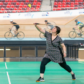 Femme en mouvement portant la robe tunique sportwear Cranberry, jouant au badminton, sur fond de cyclistes en indoor.