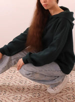 Femme portant SWEAT À CAPUCHE VERNA- -taille de 30 à 58 EU, patron de couture pdf à télécharger, en molleton vert foncé.
