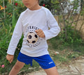 Enfant en t-shirt blanc avec motif football, jouant dehors sur pelouse, et portant un short bleu.