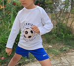 Enfant en t-shirt blanc avec motif football, jouant dehors sur pelouse, et portant un short bleu.