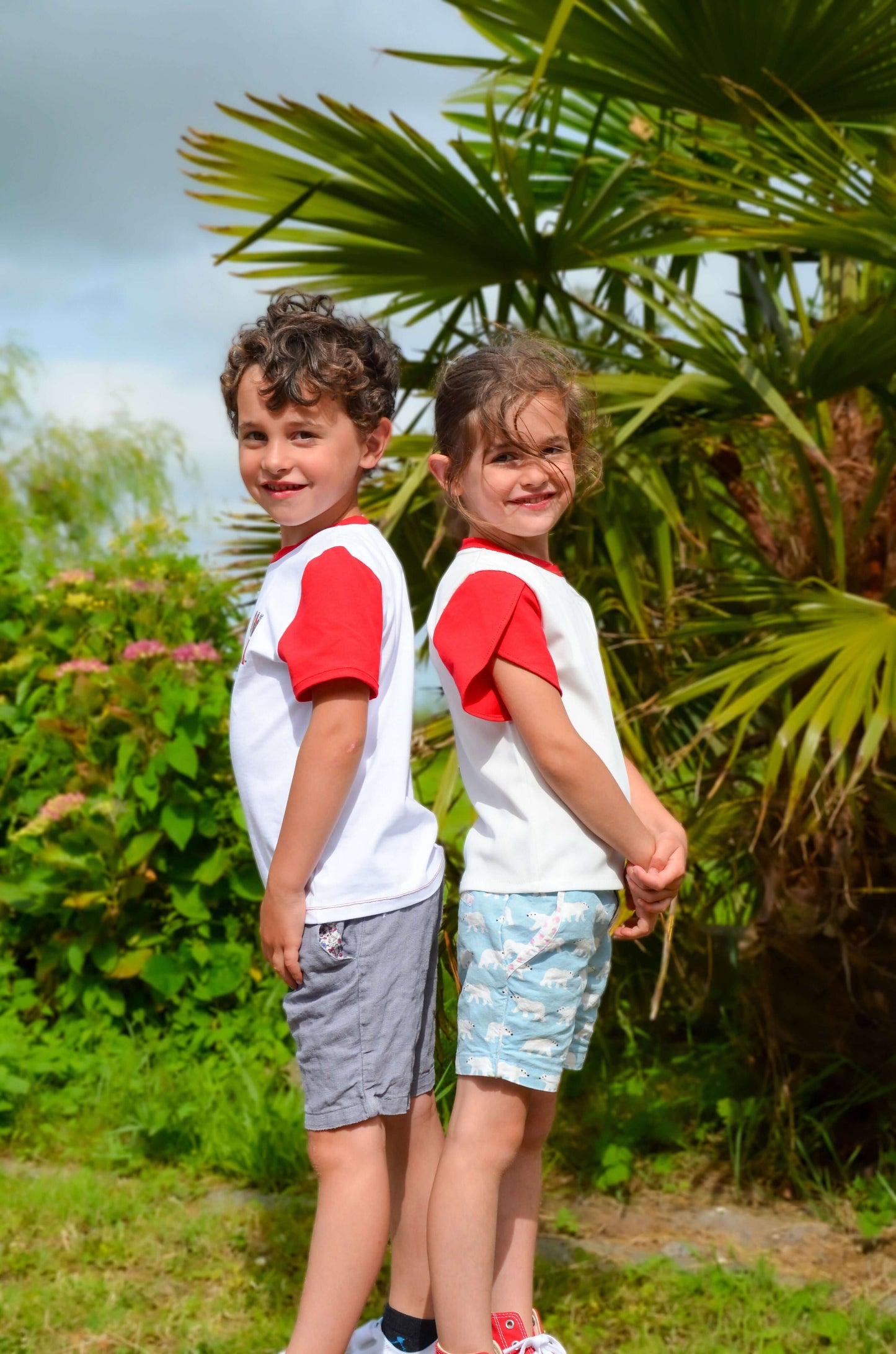 Deux enfants souriants portant des t-shirts à manches courtes rouges dans un jardin verdoyant.