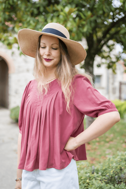 Femme souriante portant une blouse rosée et un chapeau, en plein air, avec des arbres en arrière-plan.