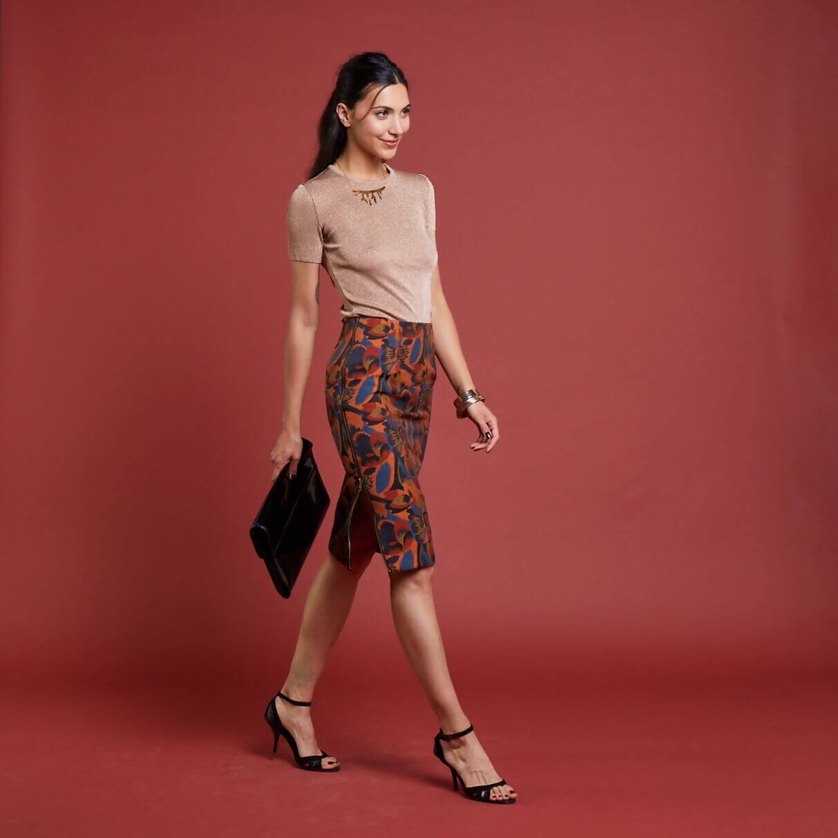 Femme élégante portant la jupe L'Ambitieuse du projet Couture pour les fêtes, marchant avec un sac à main sur fond rouge.