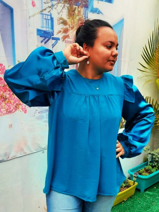Femme portant une blouse Idunn bleu avec volants aux épaules, posant dans un décor floral.