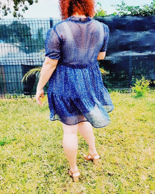 Femme portant une robe babydoll en dentelle bleu, vue de dos, dans un jardin verdoyant.