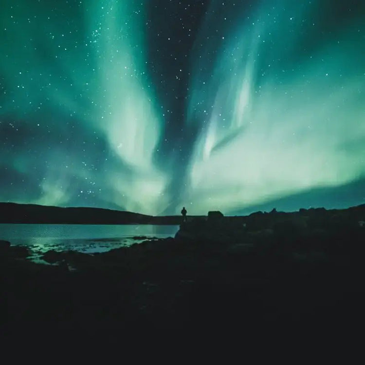 Aurore boréale illuminant le ciel étoilé au-dessus d'un paysage côtier sombre, une silhouette contemplant l'horizon lumineux.