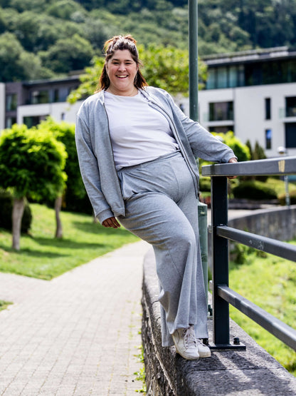 Personne souriante portant un ensemble décontracté gris avec un fond de bâtiment et verdure en arrière-plan.