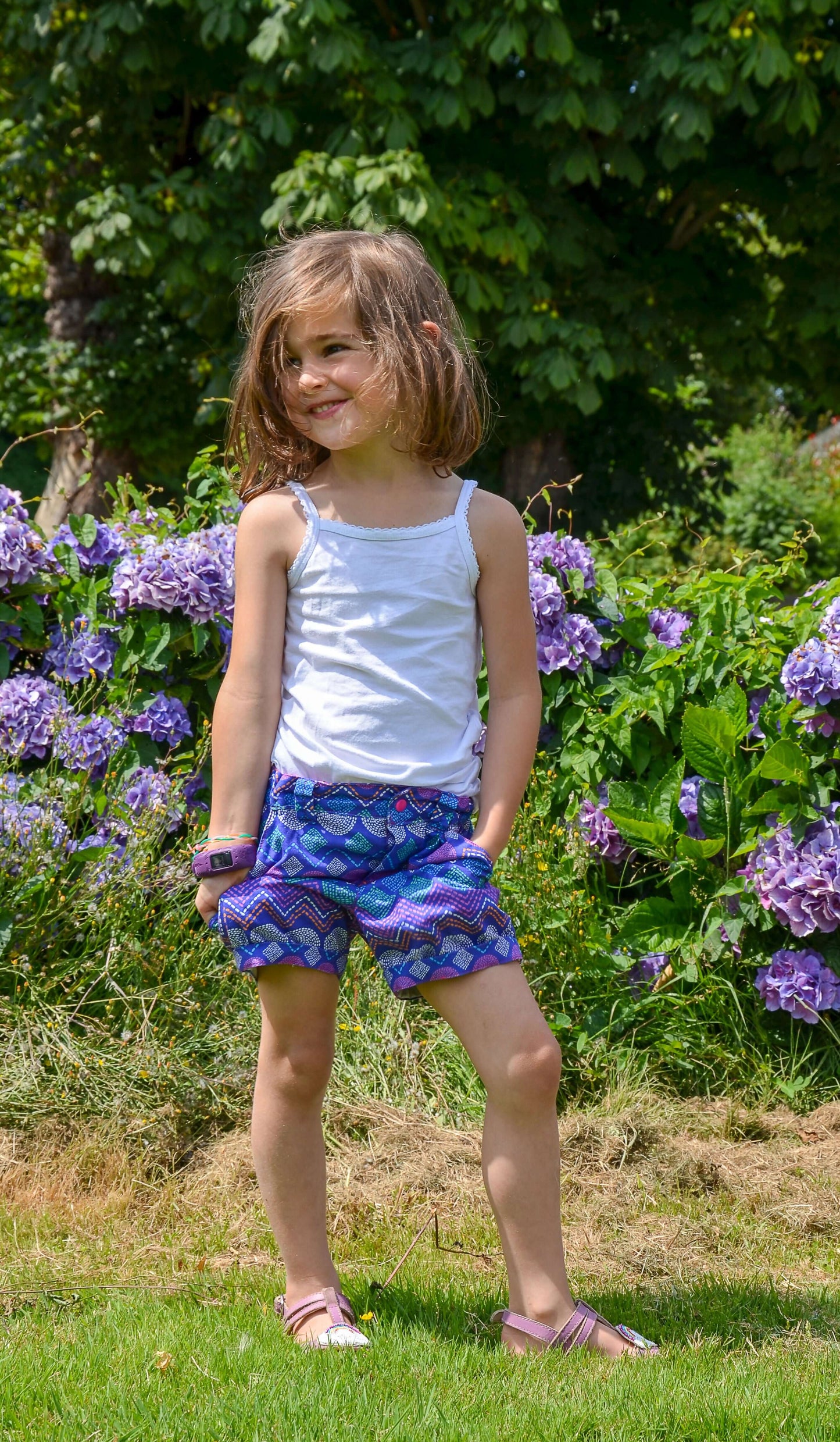 Jeune fille souriante avec un débardeur blanc et un bermuda coloré, posant dans un jardin fleuri.