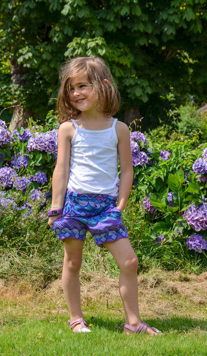 Jeune fille souriante avec un débardeur blanc et un bermuda coloré, posant dans un jardin fleuri.
