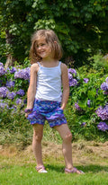 Jeune fille souriante avec un débardeur blanc et un bermuda coloré, posant dans un jardin fleuri.