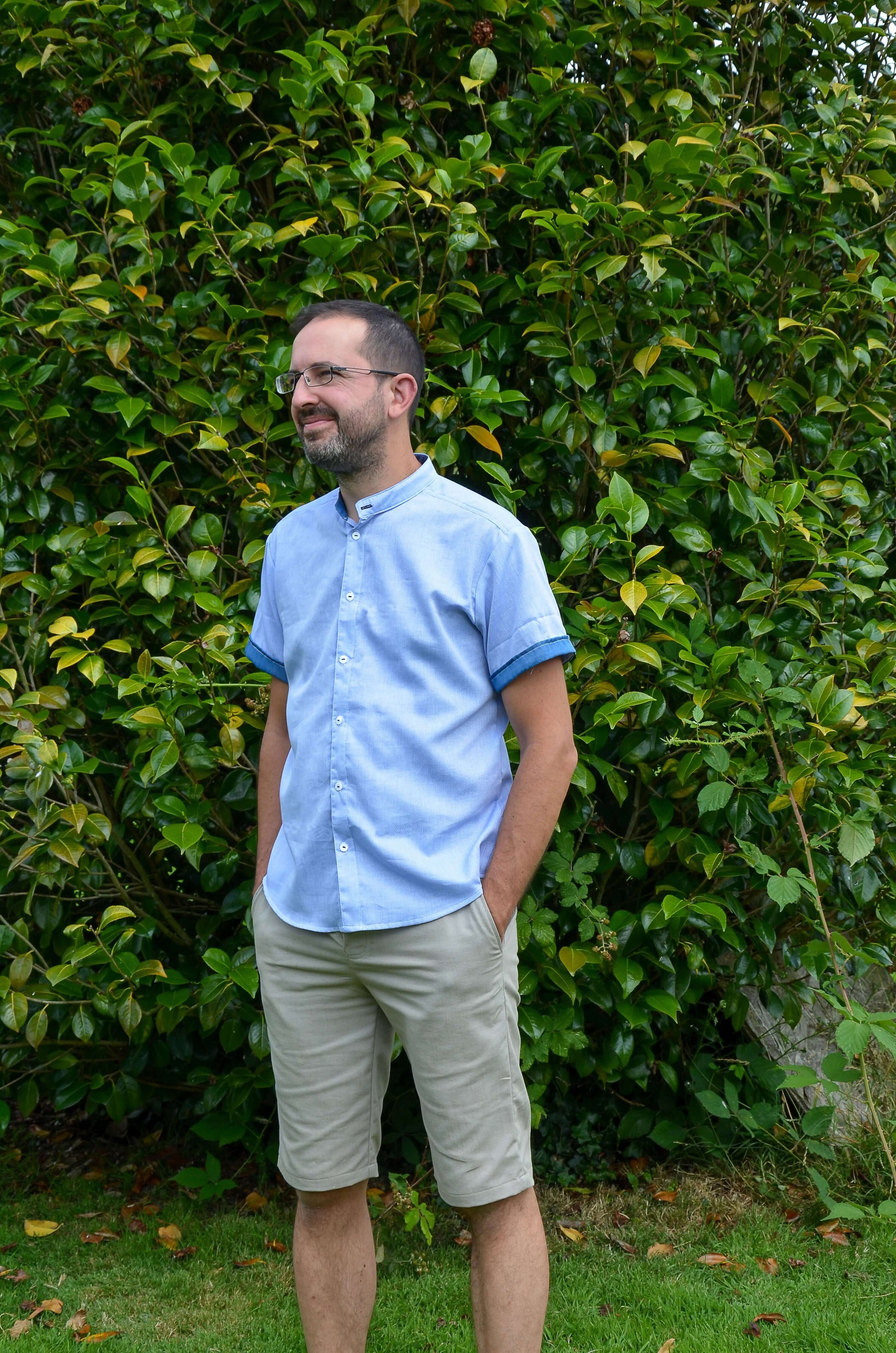 Homme portant une chemise bleue avec shorts beige devant un feuillage verdoyant, look décontracté pour l'été.