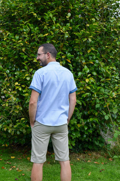 Homme de dos en chemise bleue et bermuda tendance, avec une verdure luxuriante en arrière-plan.