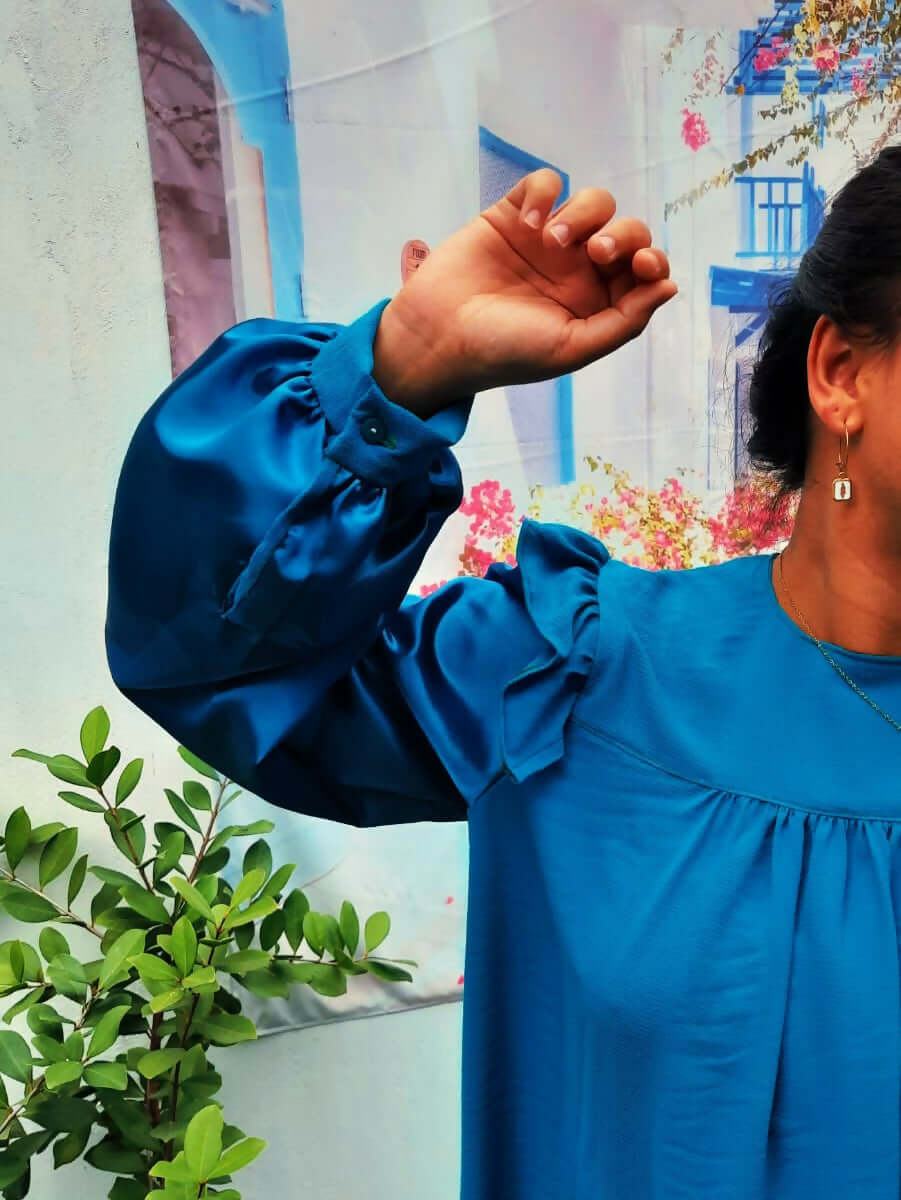 Blouse Idunn bleu avec volants aux épaules, manches Bishop, portée devant un mur coloré.
