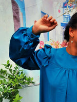 Blouse Idunn bleu avec volants aux épaules, manches Bishop, portée devant un mur coloré.