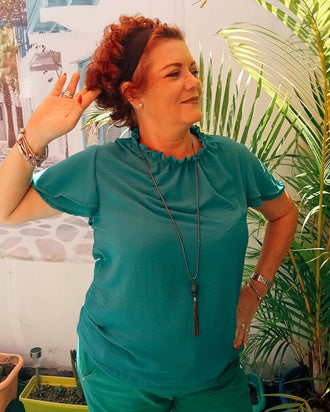 Femme portant une blouse Némésis en turquoise avec col froncé, se tenant devant des plantes en été.