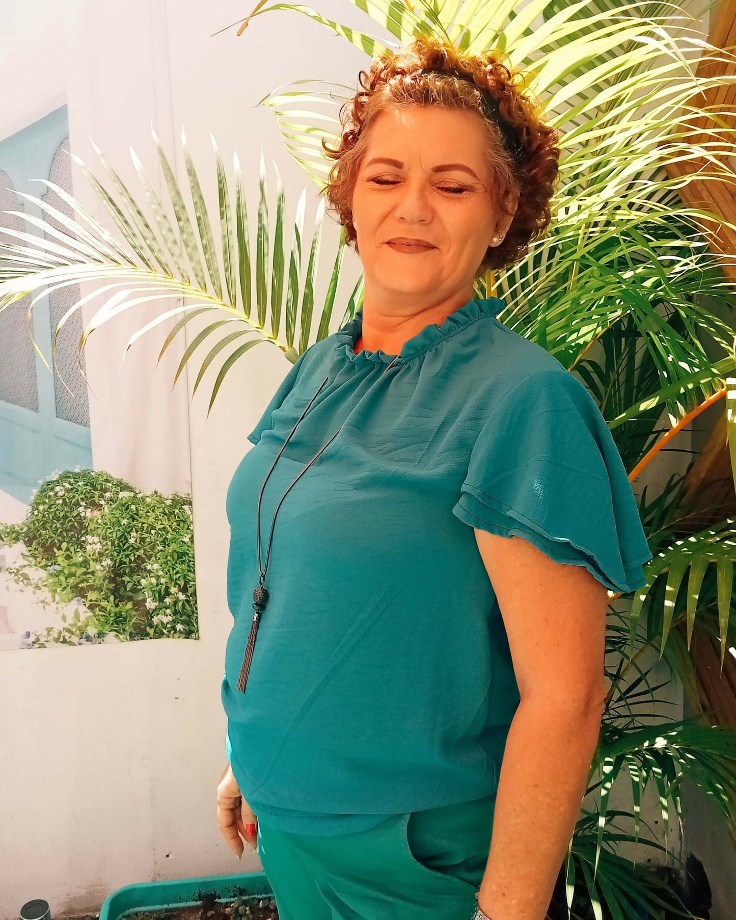 Femme souriante portant une blouse turquoise à col montant et manches volantées, devant des plantes vertes.