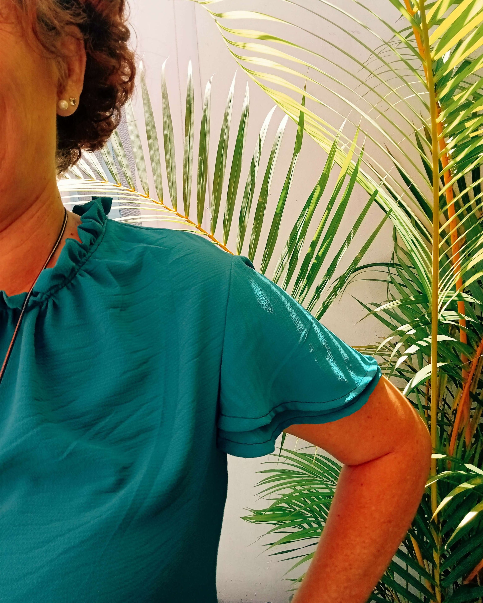 Femme portant une blouse Némésis turquoise avec col montant froncé, devant une plante tropicale.