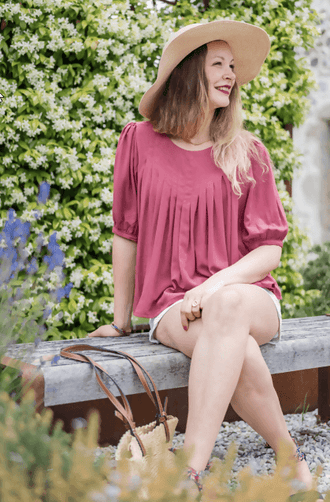 Femme souriante portant la blouse ROSAE, assise sur un banc entourée de fleurs, avec un chapeau et des shorts.