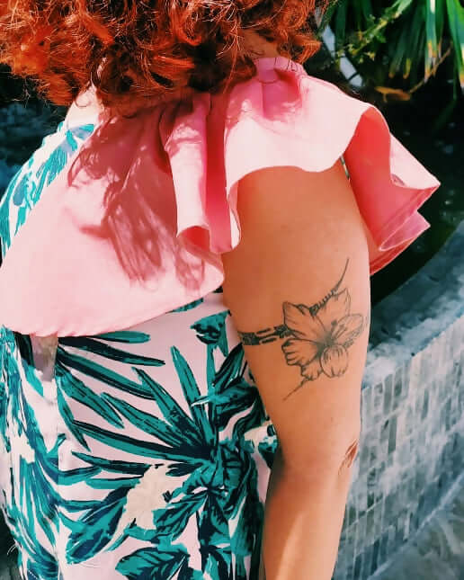 Femme avec blouse asymétrique rose et tatouage floral sur le bras, entourée de verdure.