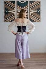 Femme portant une blouse avec corset noir et pantalon lilas, vue de dos, devant des tableaux géométriques décoratifs.