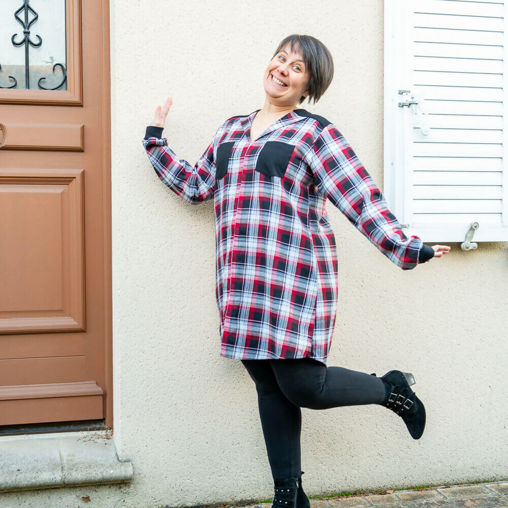 Femme souriante en blouse rouge à carreaux, style chemise, posant devant une porte.