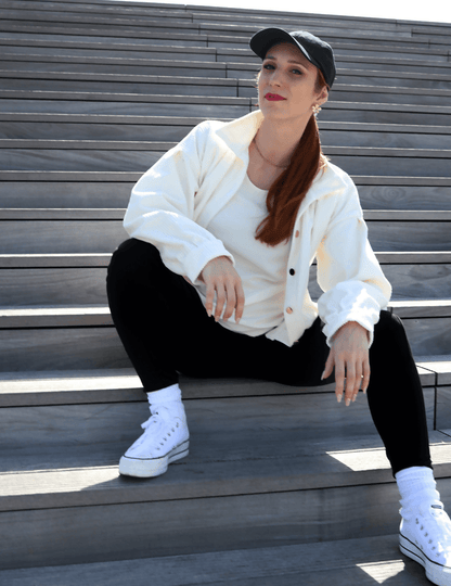 Femme décontractée portant un blouson Cosmos blanc, assise sur des marches en bois sous un soleil brillant.