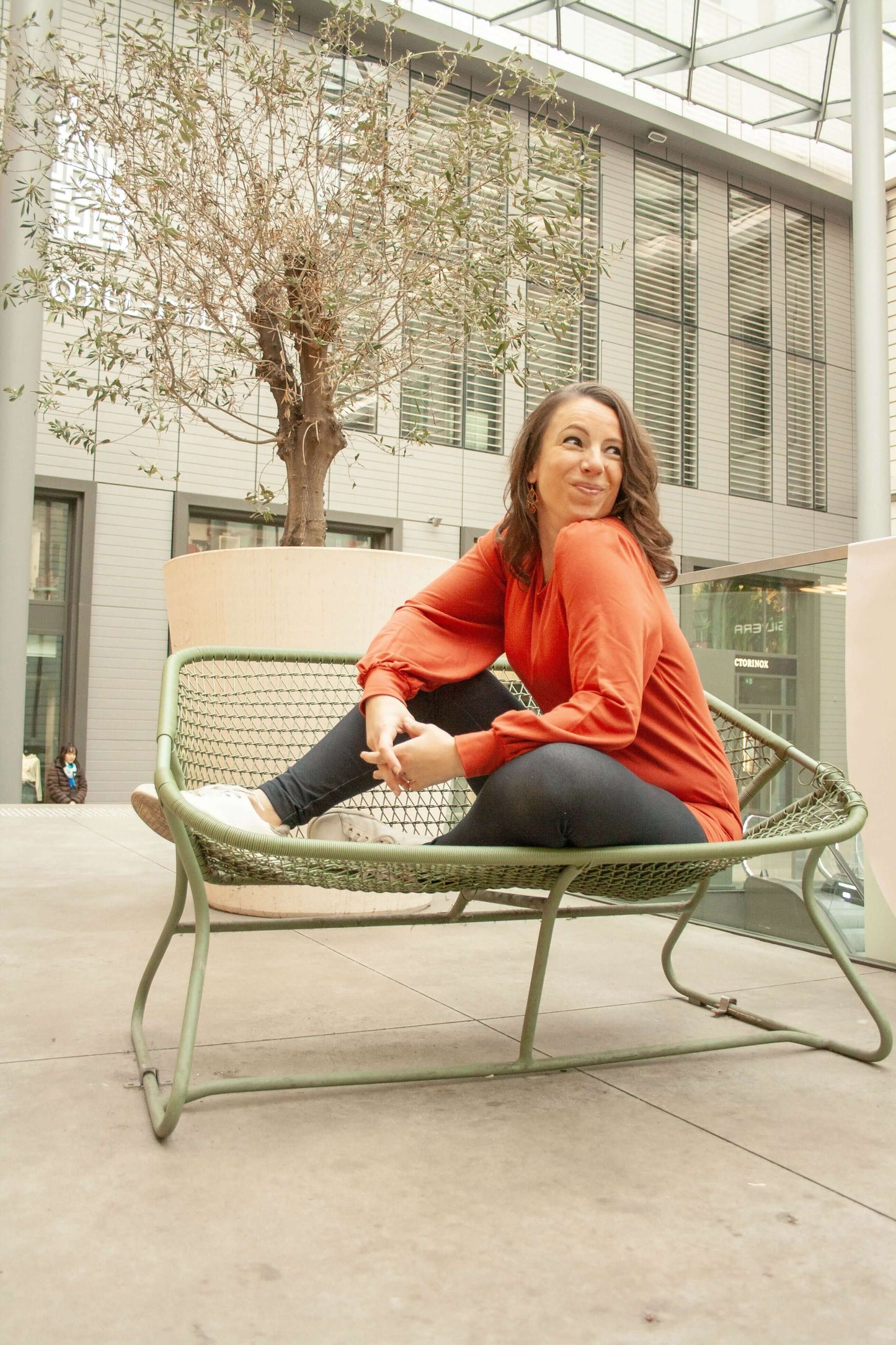 Femme assise sur un banc en métal dans un espace urbain moderne, portant un pull orange vif.
